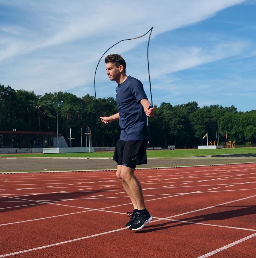 Homme sautant à la corde sur une piste rouge. Corde noire, poignées bleues.