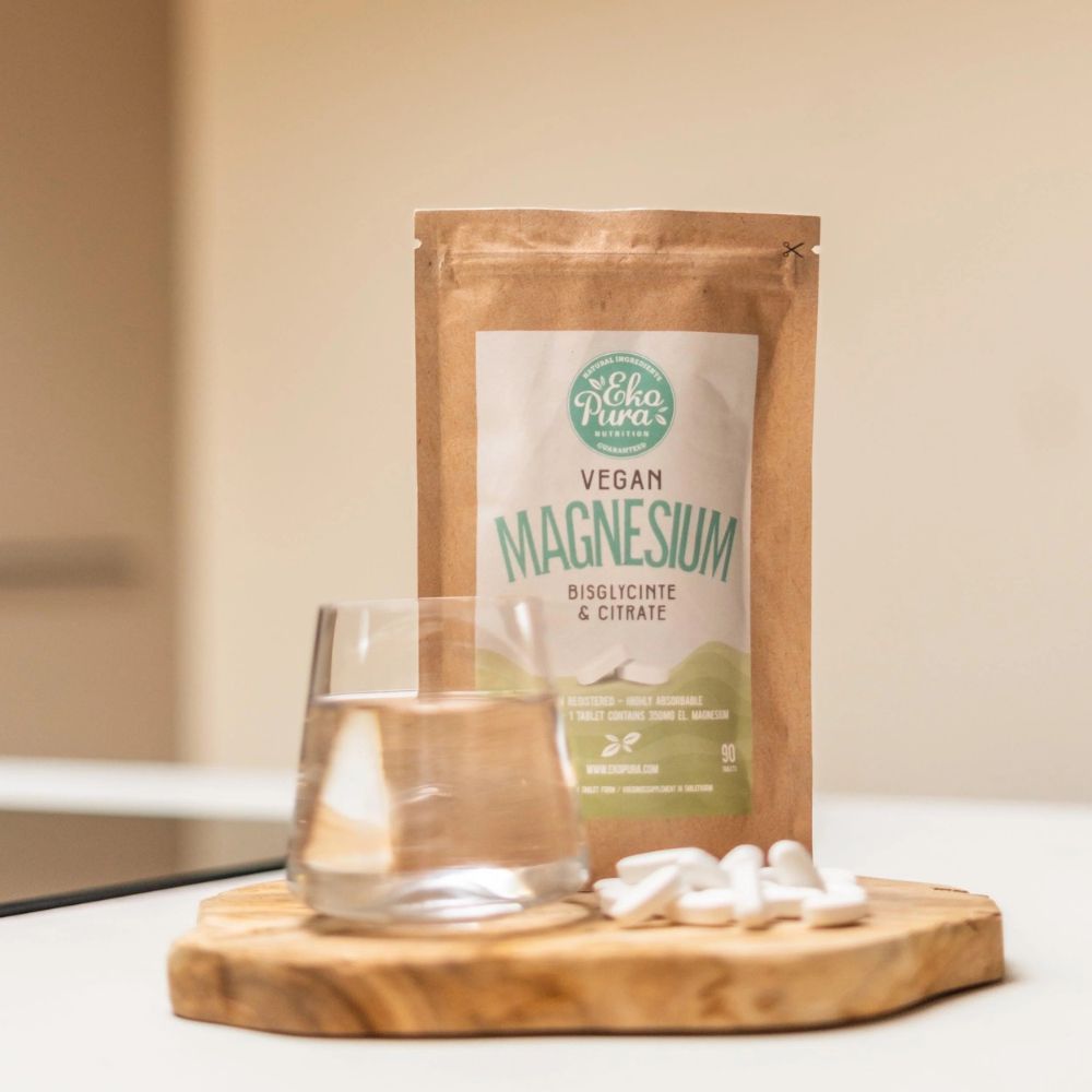Bruine verpakking met wit etiket. Op het etiket staat "Vegan Magnesium". Daarnaast een glas water en tabletten.