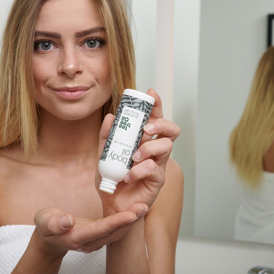 Vrouw houdt een kleine fles Body Oil vast. Witte fles met grijze bladeren. Tekst: Tea Tree Oil, Australian Bodycare.