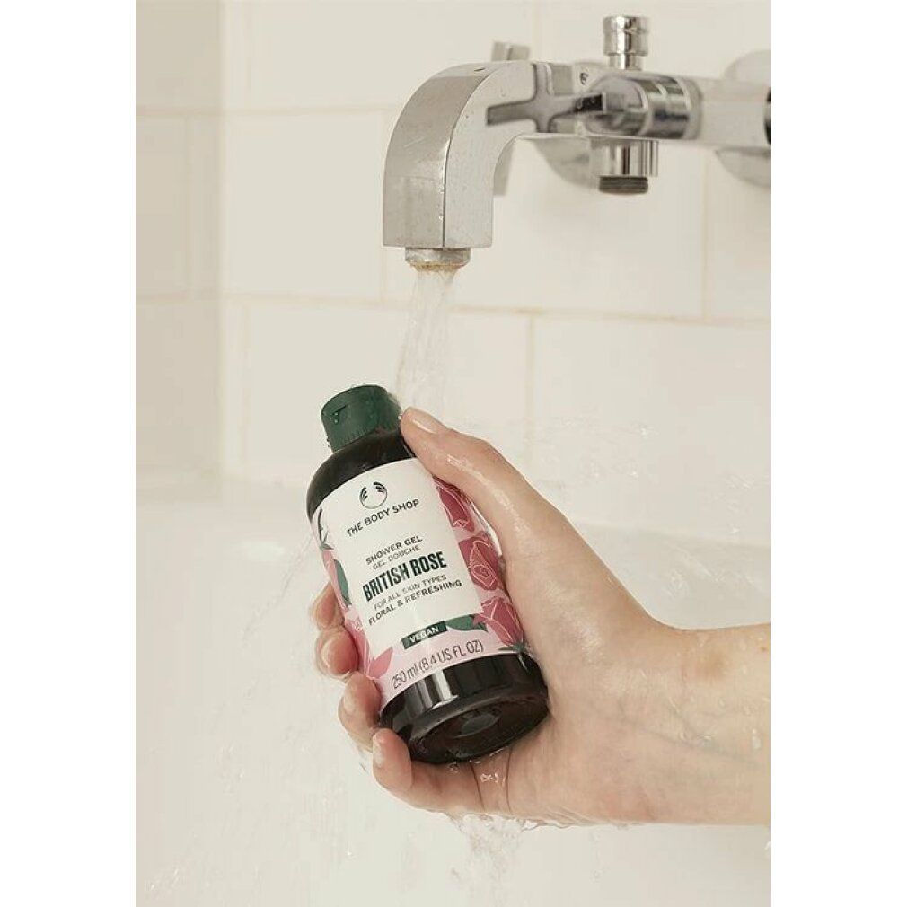 Main tenant un flacon de gel douche British Rose sous un jet d'eau. Bouchon vert, étiquette blanche avec motif floral. Vegan.