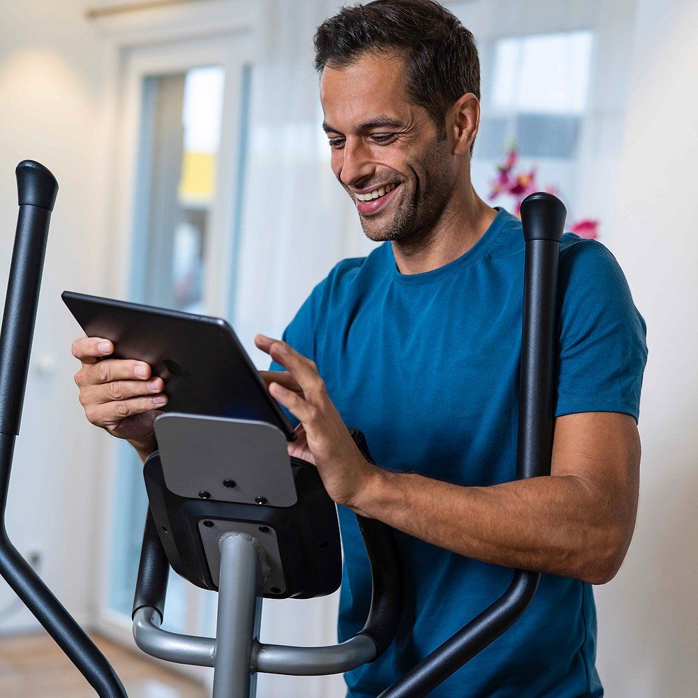 Personne utilise une tablette sur un crosstrainer. Support tablette. Panneau noir. La personne sourit. Arrière-plan : pièce.