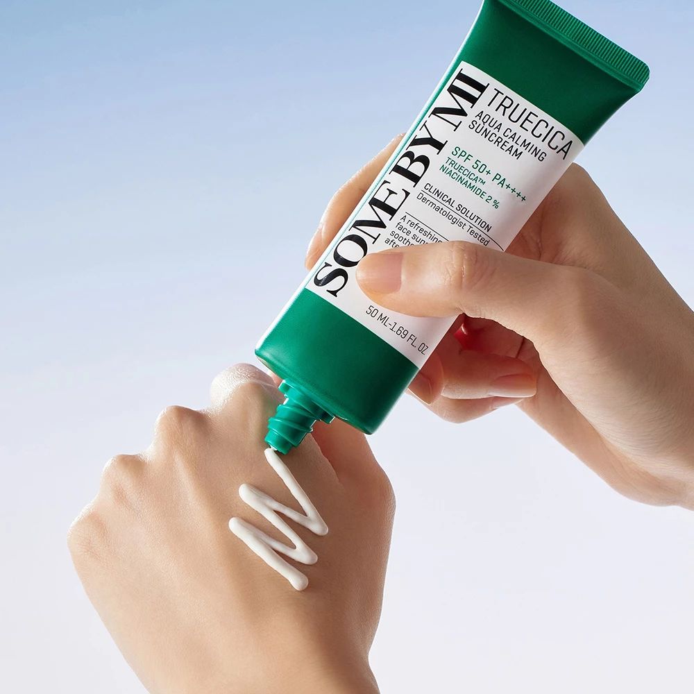 Main pressant de la crème solaire d'un tube. Texte : SOMEBYMI, TRUECICA Aqua Calming Suncream, SPF 50+ PA++++. Crème sur la peau.