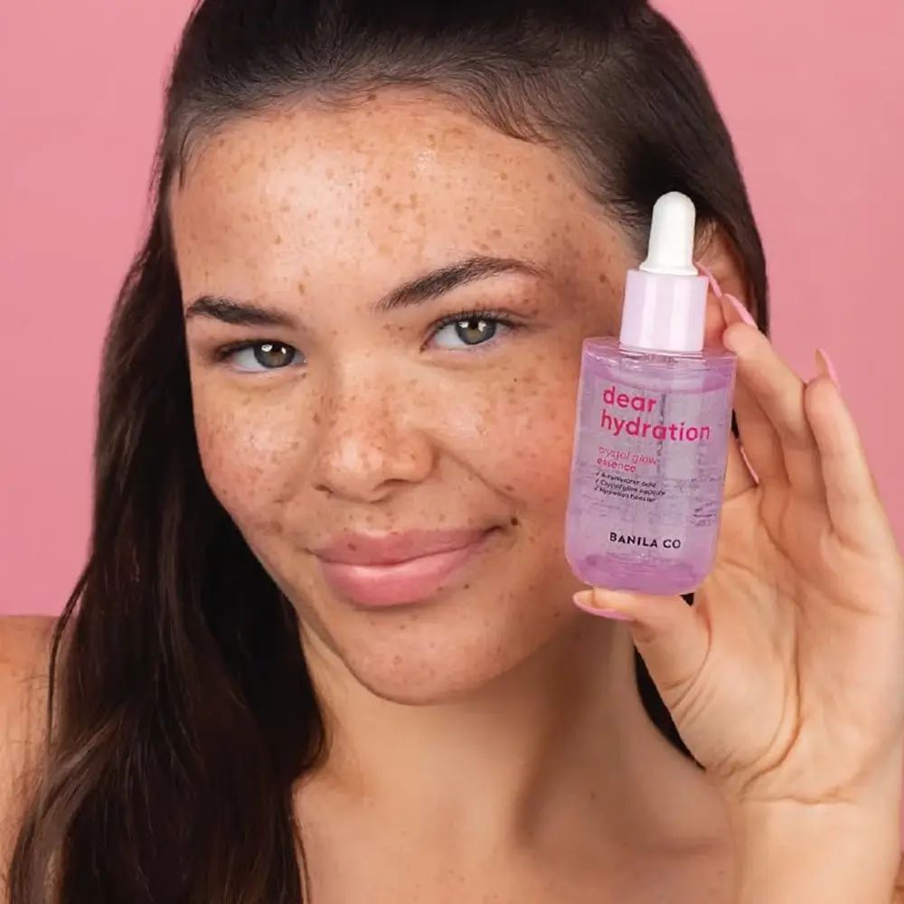 Vrouw houdt flesje BANILA CO Dear Hydration Crystal Glow Essence vast. Roze fles met witte dop en pipet.