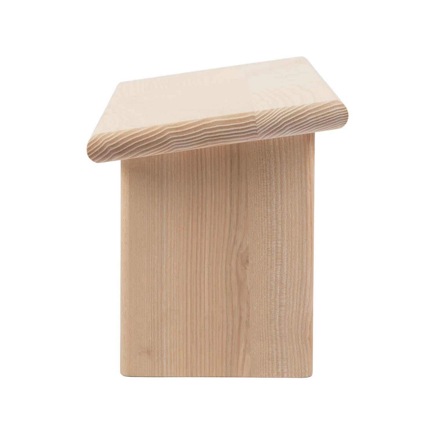 Tabouret en bois clair, vue latérale. Assise rectangulaire, deux pieds. Bords arrondis.