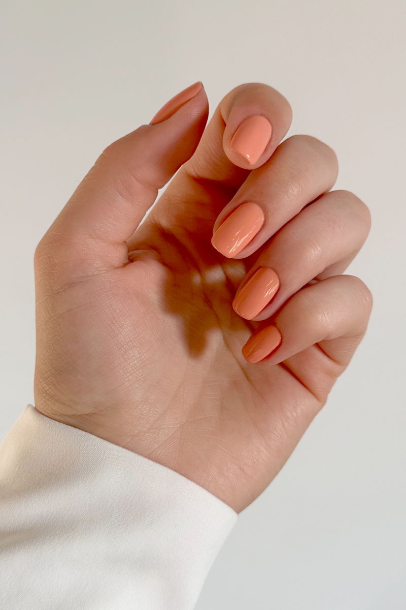 Main avec ongles vernis couleur pêche. Les ongles sont courts et de forme ovale. Le fond est blanc.