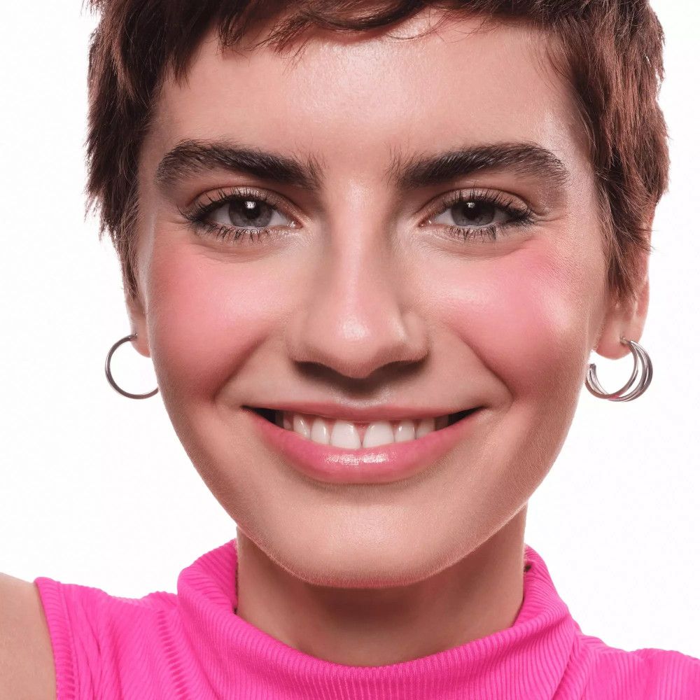 Portrait d'une femme avec du blush rose sur les joues. Elle porte des créoles argentées et un haut rose.