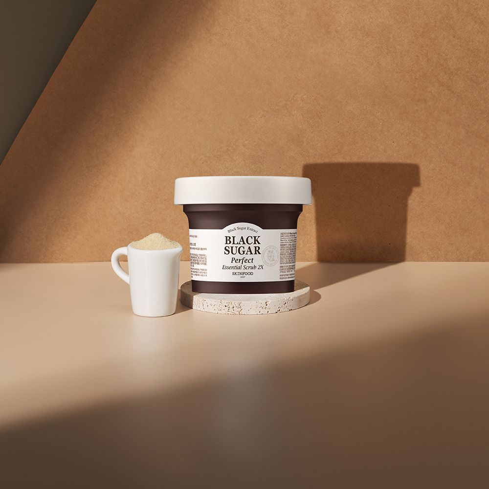 Product op een sokkel met een klein kopje ernaast. Opschrift: BLACK SUGAR Perfect Essential Scrub 2X. SKINFOOD logo.