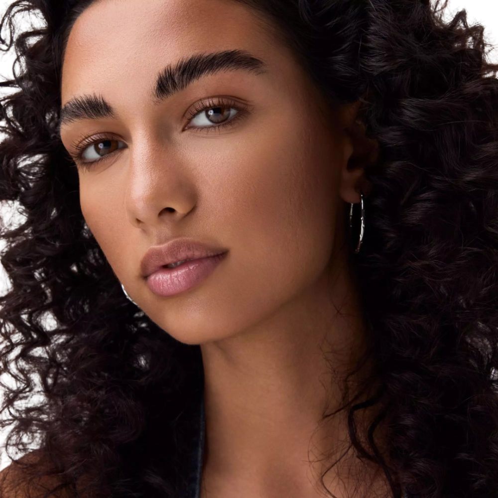 Portrait d'une femme aux cheveux bouclés et foncés. Elle porte des créoles argentées. Son teint est bronzé.