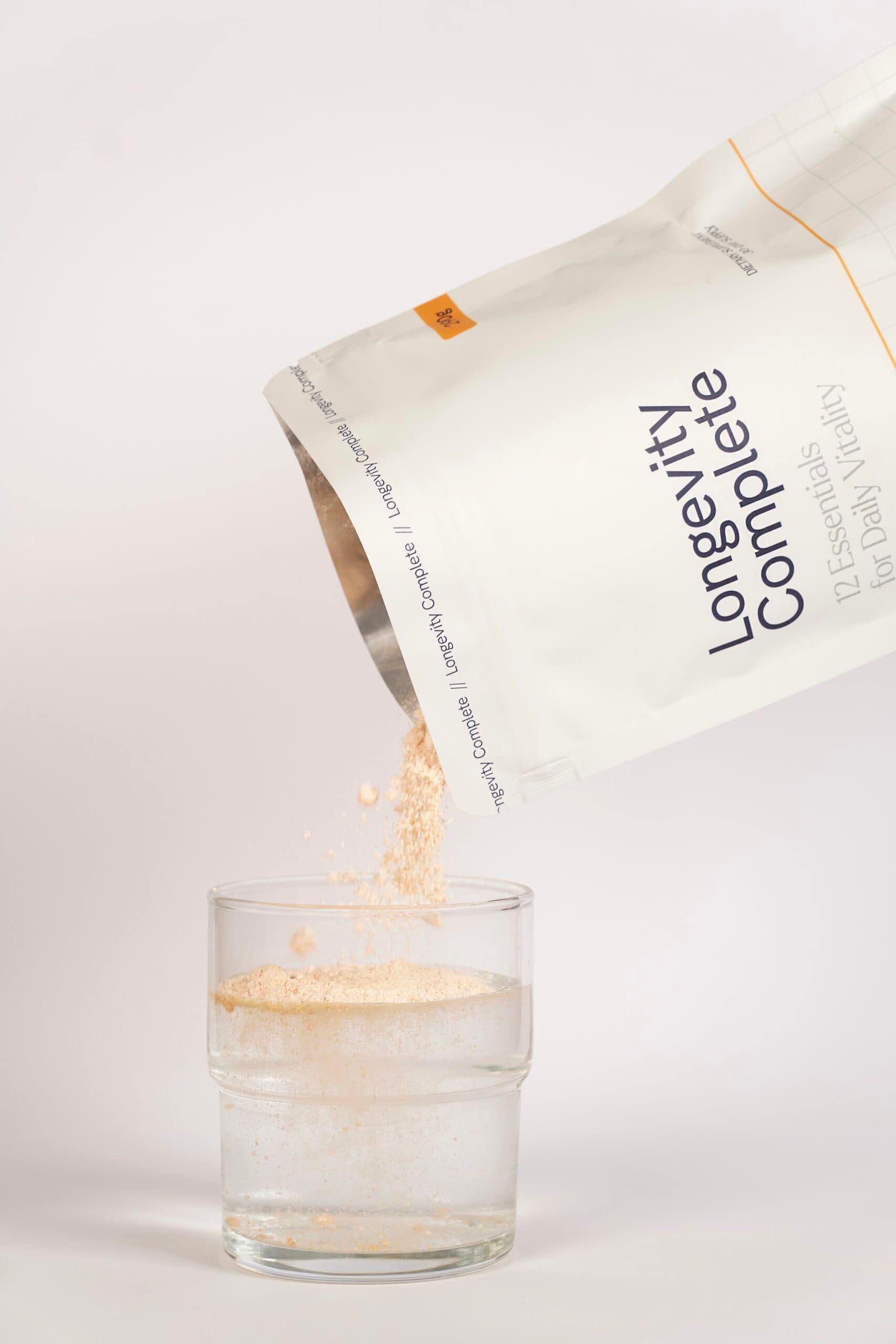 Un sachet de poudre est versé dans un verre d'eau. Le sachet est étiqueté 'Longevity Complete' et '12 Essentials for Daily Vitality'.