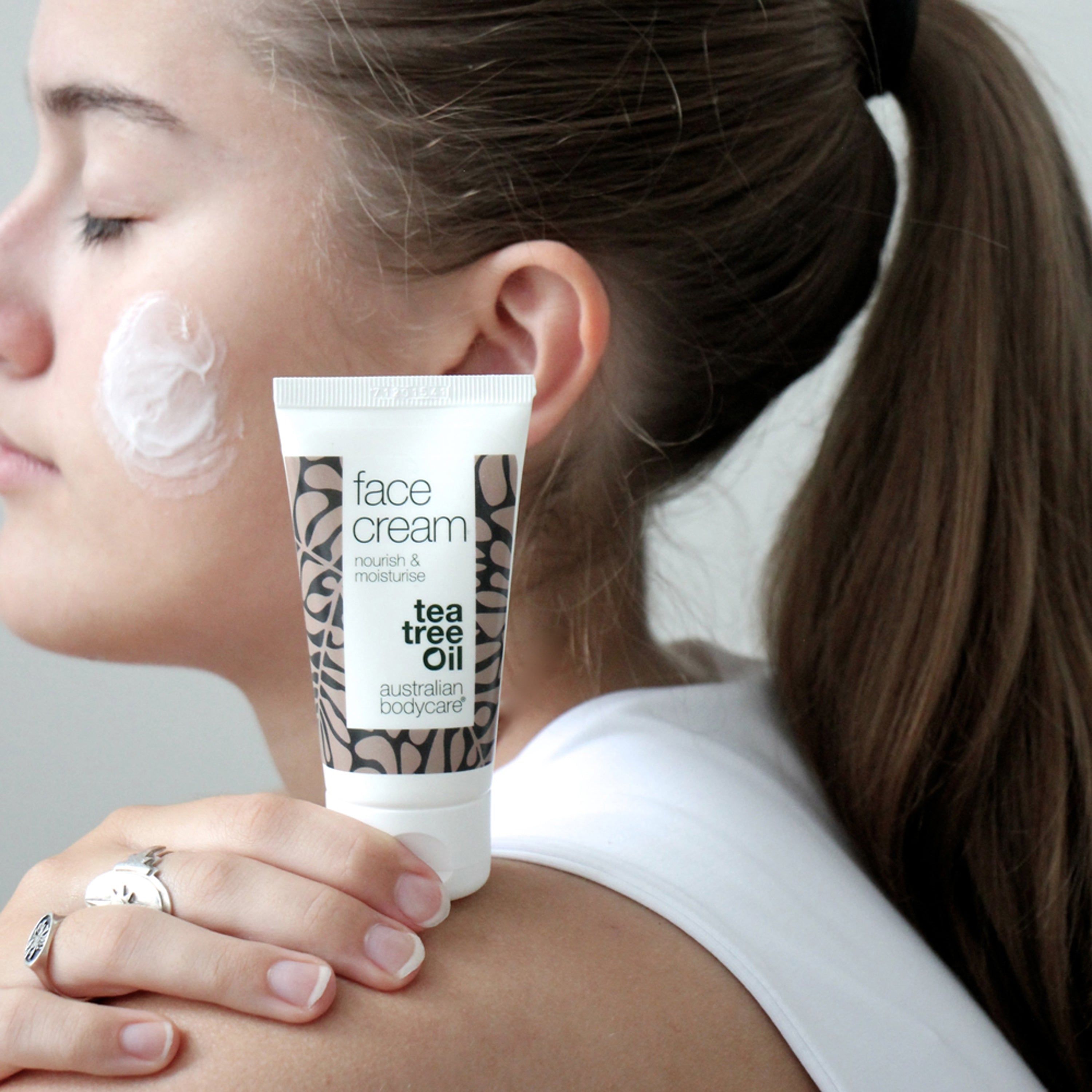 Femme avec crème sur la joue. Tube avec texte : face cream, tea tree oil. Marque : Australian Bodycare.