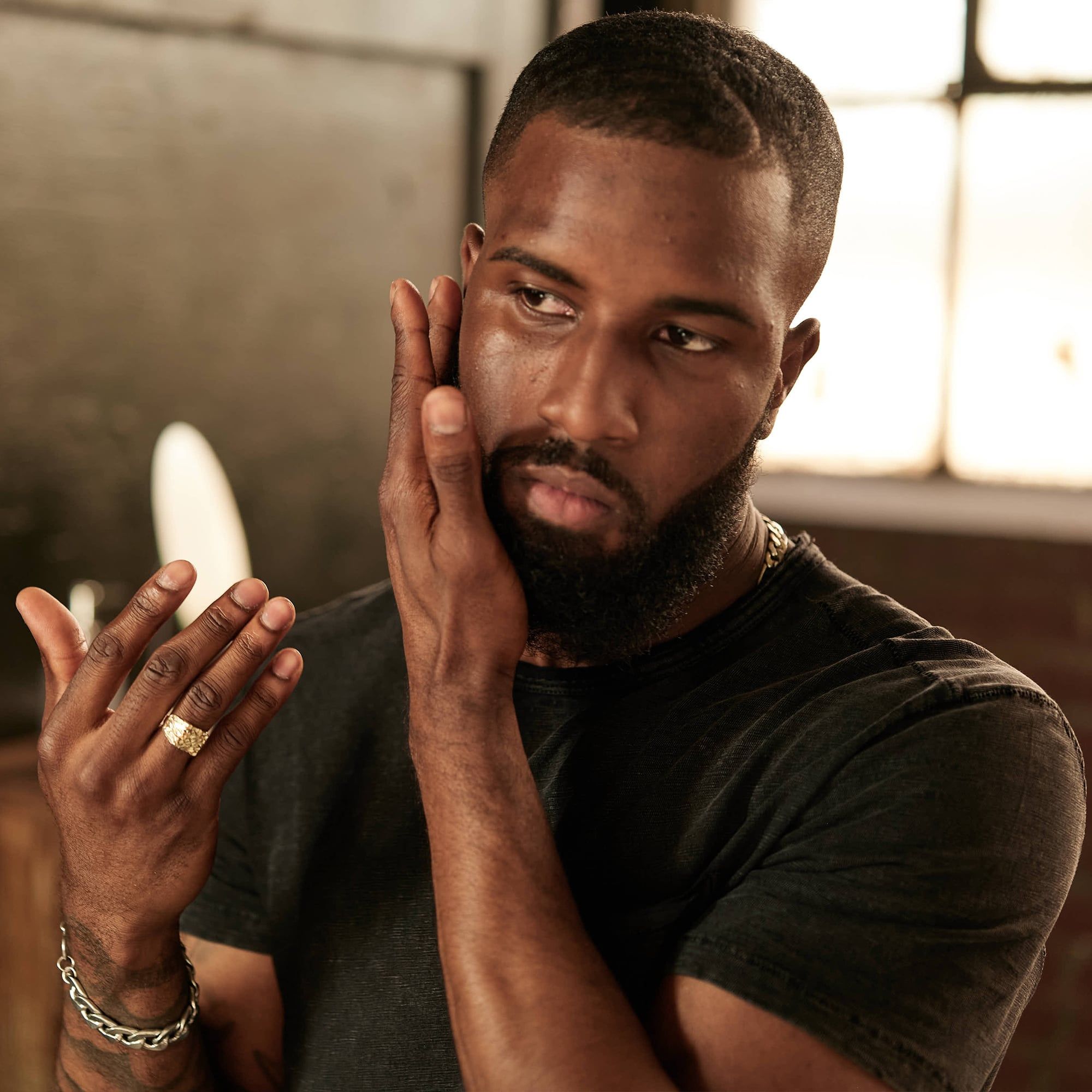 Homme barbu touchant son visage. Il porte un t-shirt noir et des bijoux. Un mur clair est visible en arrière-plan.