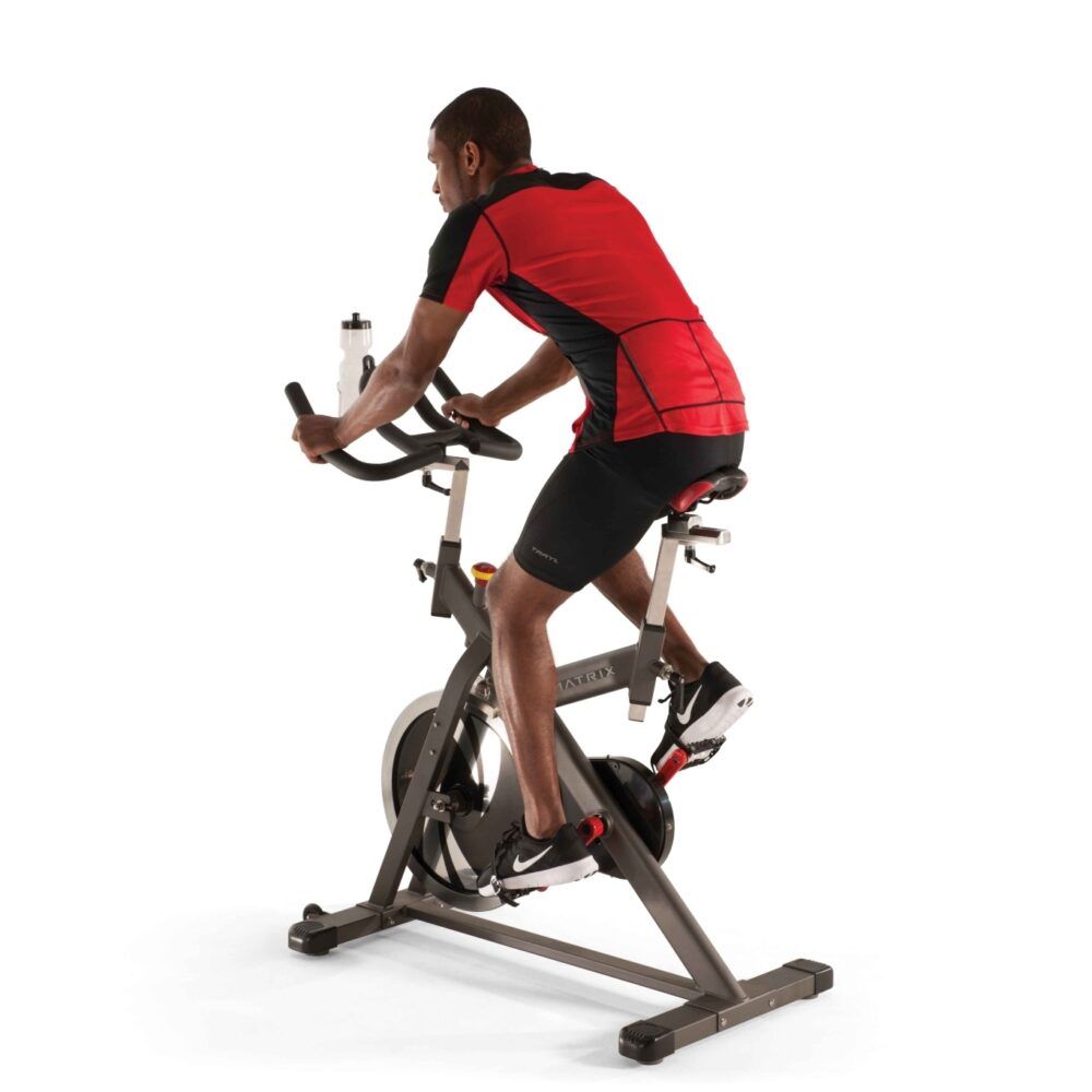 Indoor Cycle. Grijs frame, zwart stuur, rood zadel. Persoon aan het trainen. Waterfles op het stuur.