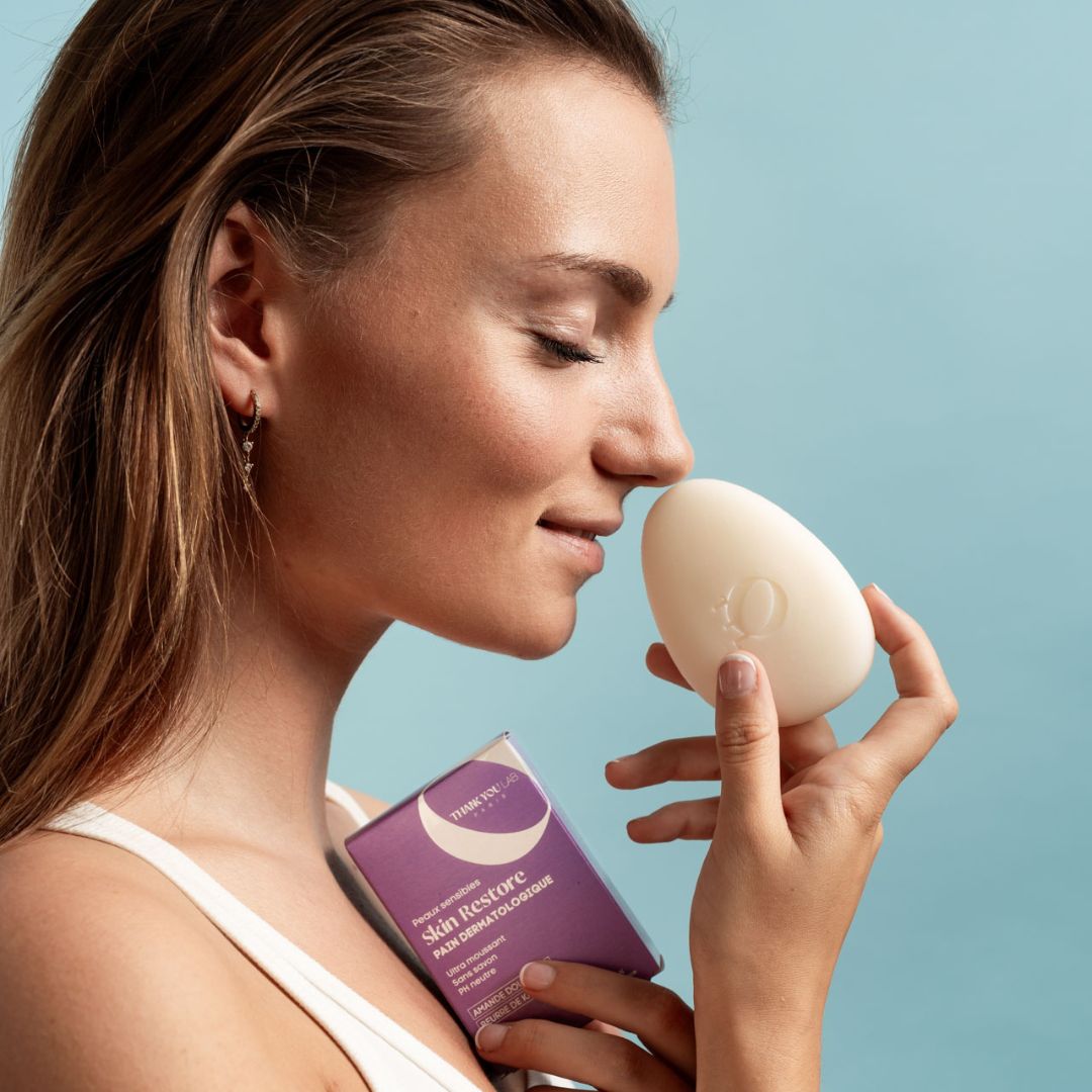 Une femme tient un pain de savon ovale et une boîte violette. Elle sent le savon.