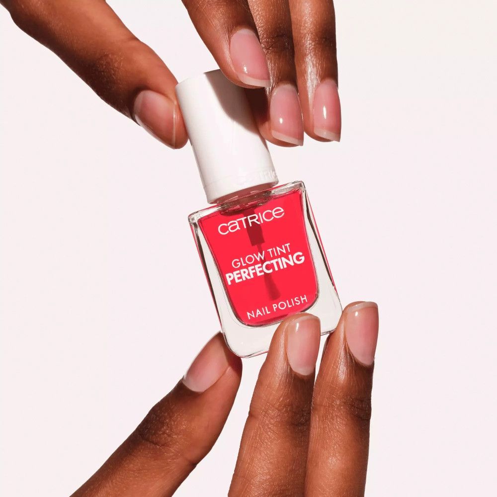 Mains tenant un flacon de vernis à ongles. Vernis rose, bouchon blanc. Inscription : Catrice, Glow Tint Perfecting, Nail Polish. Ongles vernis.