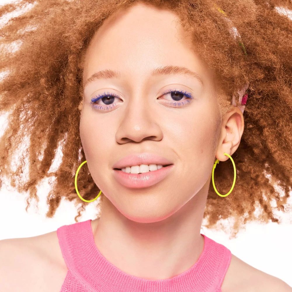 Gros plan d'une femme aux cheveux bouclés. Elle porte des boucles d'oreilles jaunes et un haut rose. Eyeliner bleu.