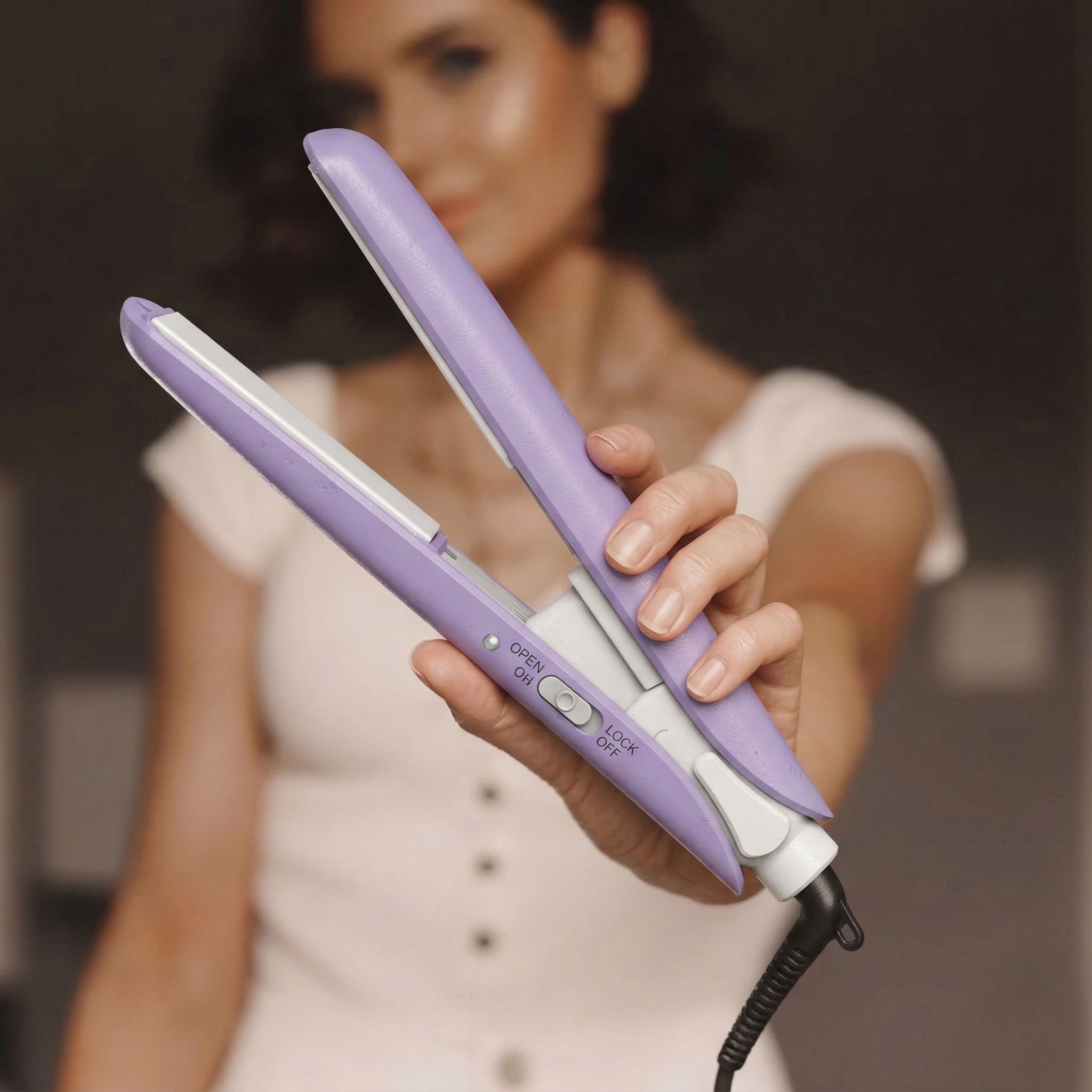 Une femme tient un lisseur violet avec un cordon blanc et des commandes visibles.