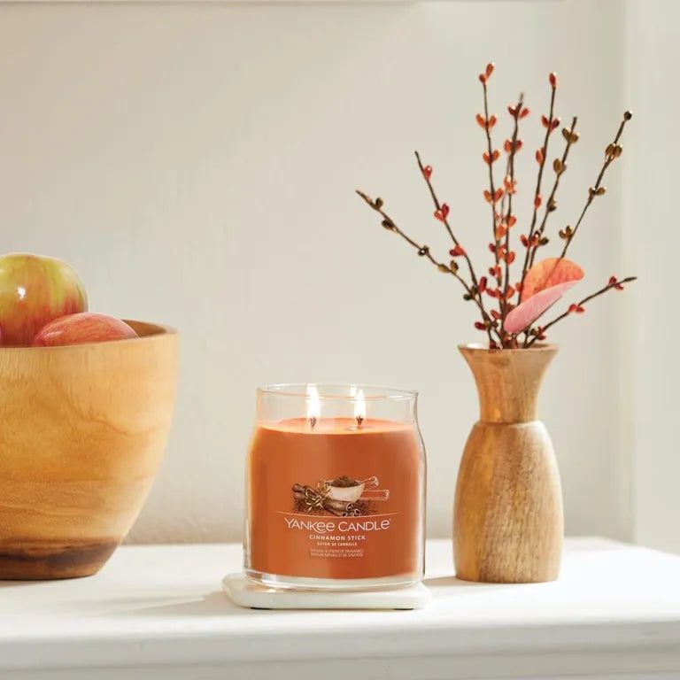 Pot en verre avec cire orange sur un dessous de plat blanc. À côté, un bol en bois avec des pommes et un vase avec des branches.