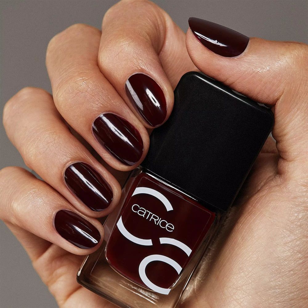 Main avec ongles vernis et flacon de vernis à ongles. Ongles bordeaux, flacon avec logo Catrice.