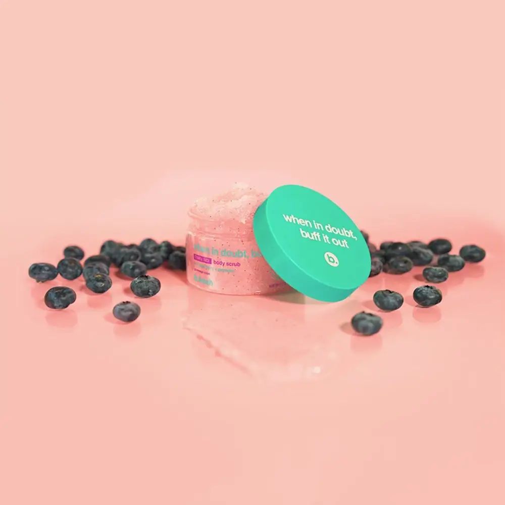 Open roze pot met lichaamsscrub en turquoise deksel. Blauwe bessen liggen rond het product.