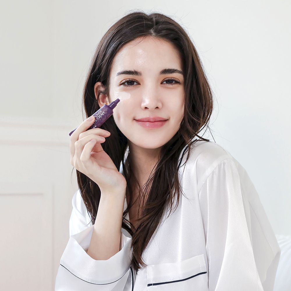 Vrouw houdt tube vast. Gedrukt: Collagen Power Firming Eye Cream. Merk MIZON. Vrouw draagt wit overhemd.