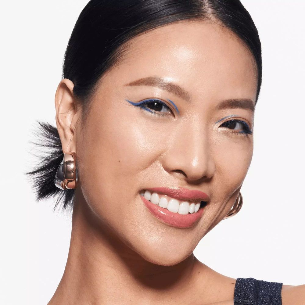 Portrait d'une femme avec un trait de khôl bleu sur l'œil. Elle porte des boucles d'oreilles et sourit.