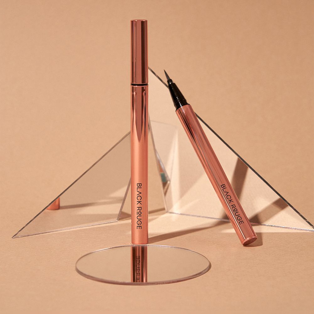 Twee zwarte eyeliner pennen met roségouden behuizing. Spiegels en een ronde basis op de achtergrond. De merknaam is zichtbaar.