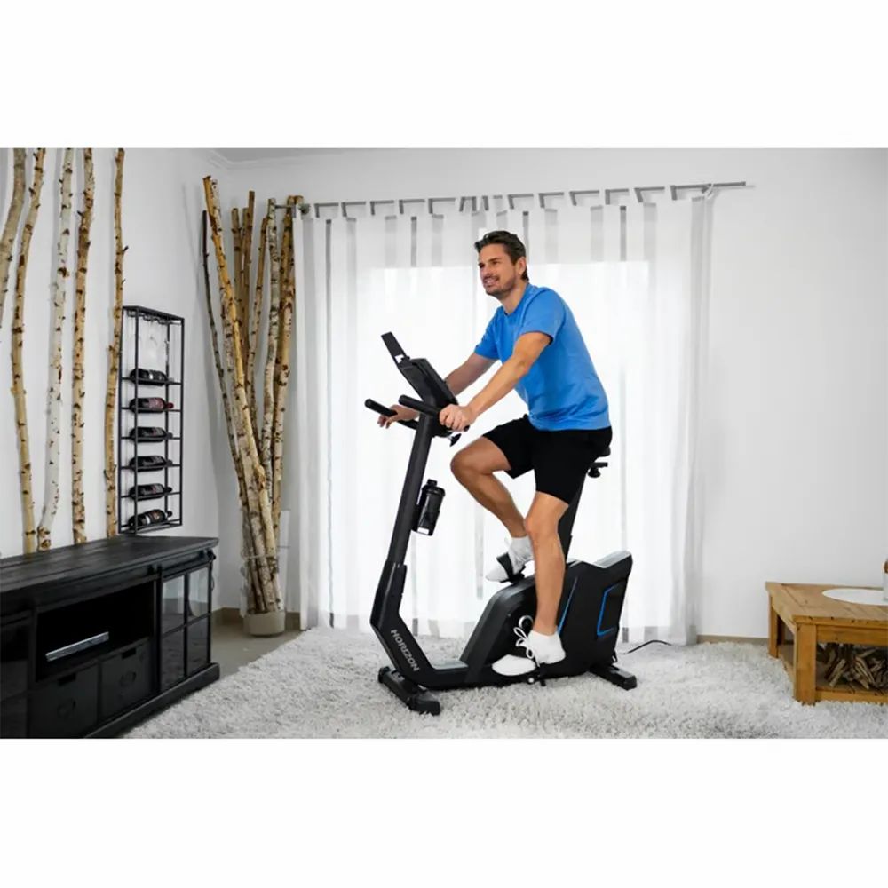 Homme sur ergomètre Horizon Fitness 5.0Ui. Dans un salon. Avec écran, pédales, selle et porte-gobelet.
