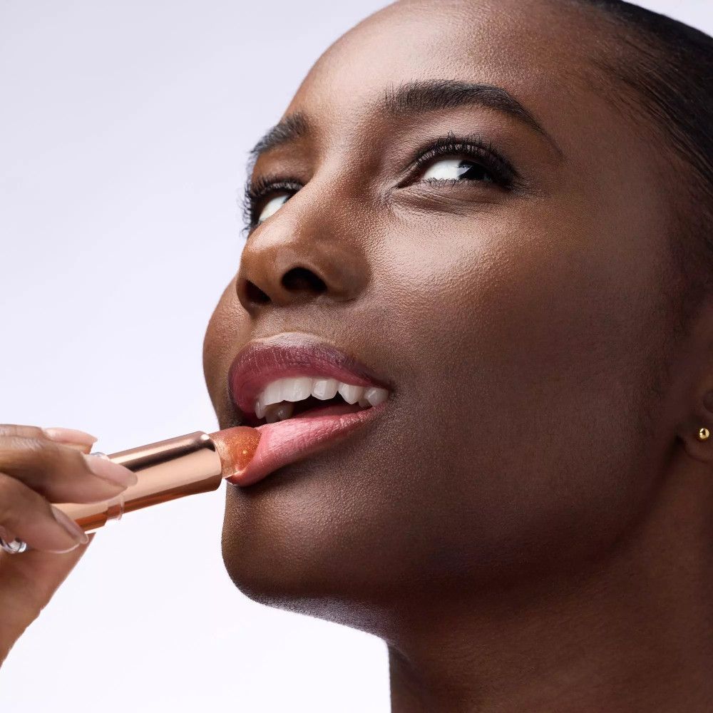 Femme à la peau foncée appliquant du baume à lèvres. Baume à lèvres or rose. Lèvres légèrement ouvertes.