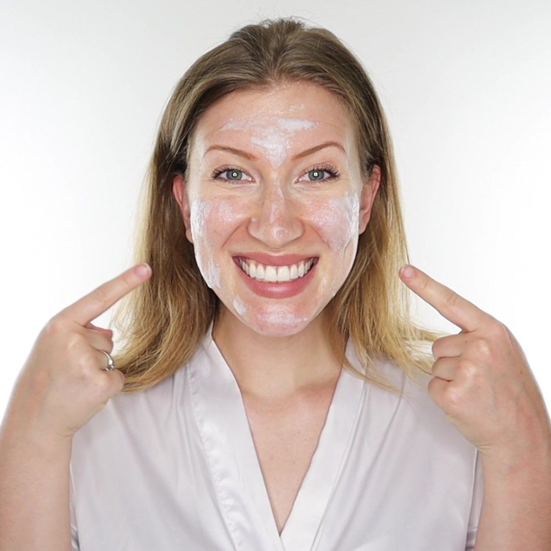 Vrouw met witte crème op haar gezicht. Ze wijst met haar vingers naar haar gezicht. Ze draagt een witte badjas.