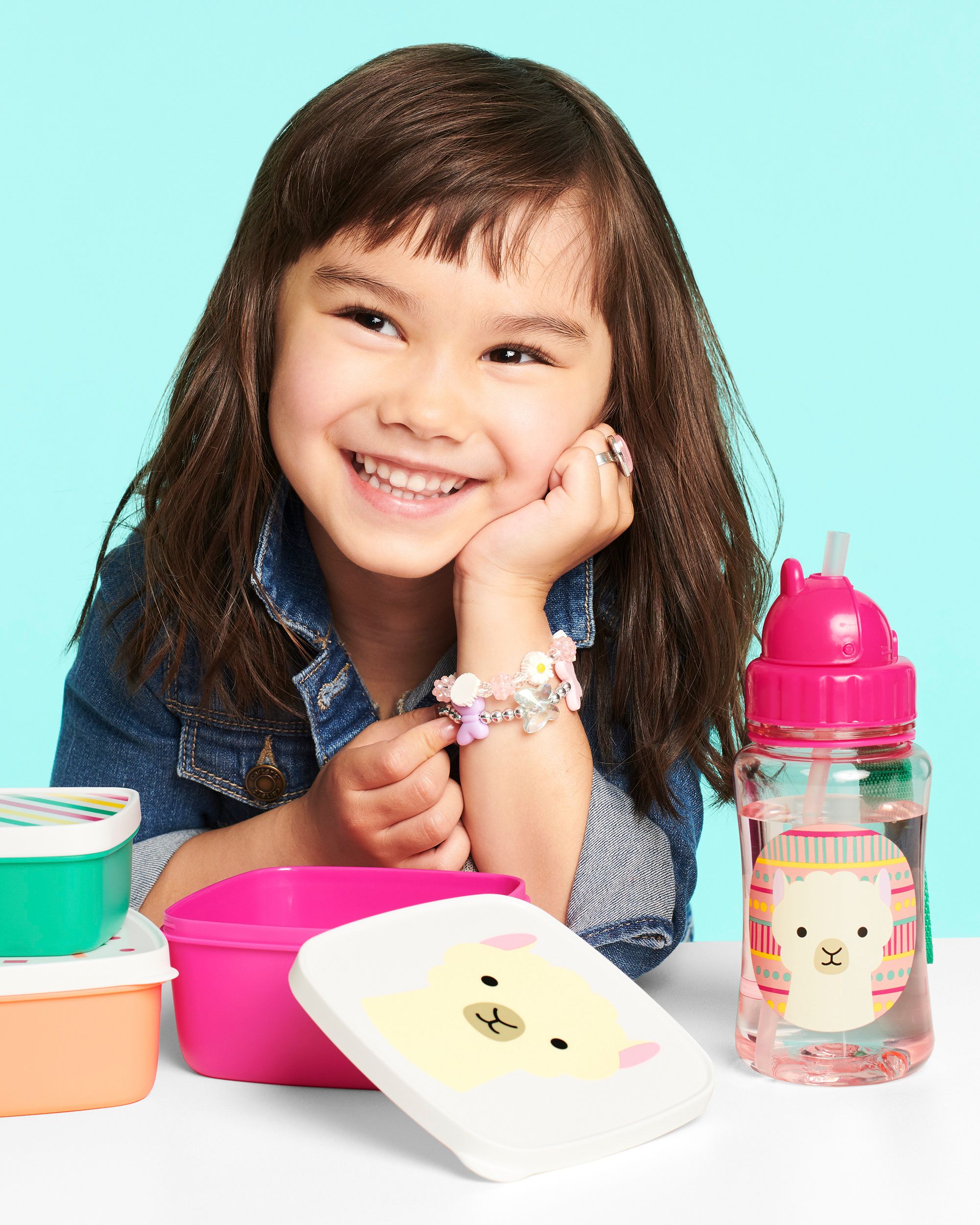 Kind met drinkfles en lunchboxen. Roze drinkfles met lama-motief. Lunchboxen in verschillende kleuren. Kind draagt spijkergoedhemd.