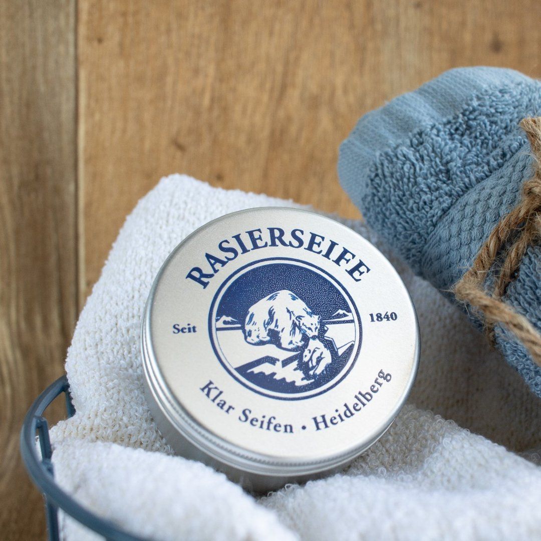 Scheerzeep in blik op handdoek. Opschrift: Rasierseife, Klar Seifen, Heidelberg. Logo met beer. Blauwe handdoekrol op de achtergrond.