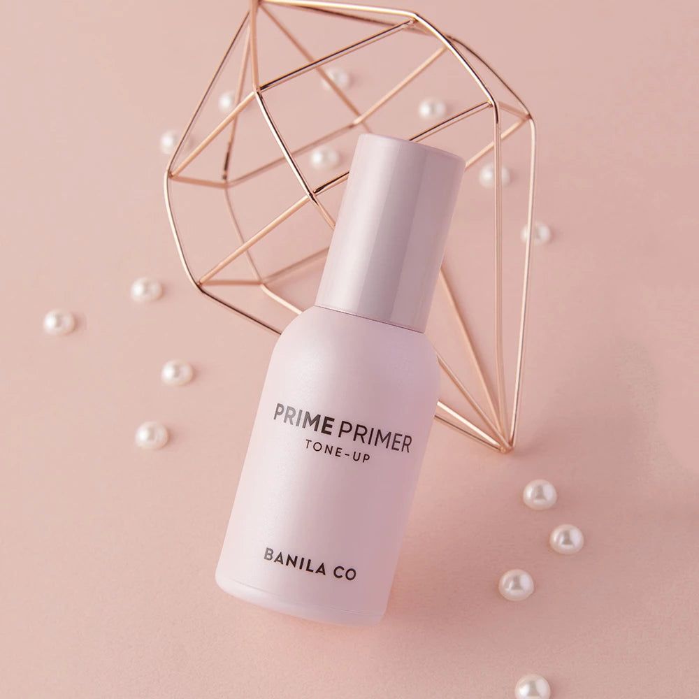 Flacon de primer rose avec bouchon. Inscription : PRIME PRIMER TONE-UP, BANILA CO. Décoration avec perles et forme dorée.