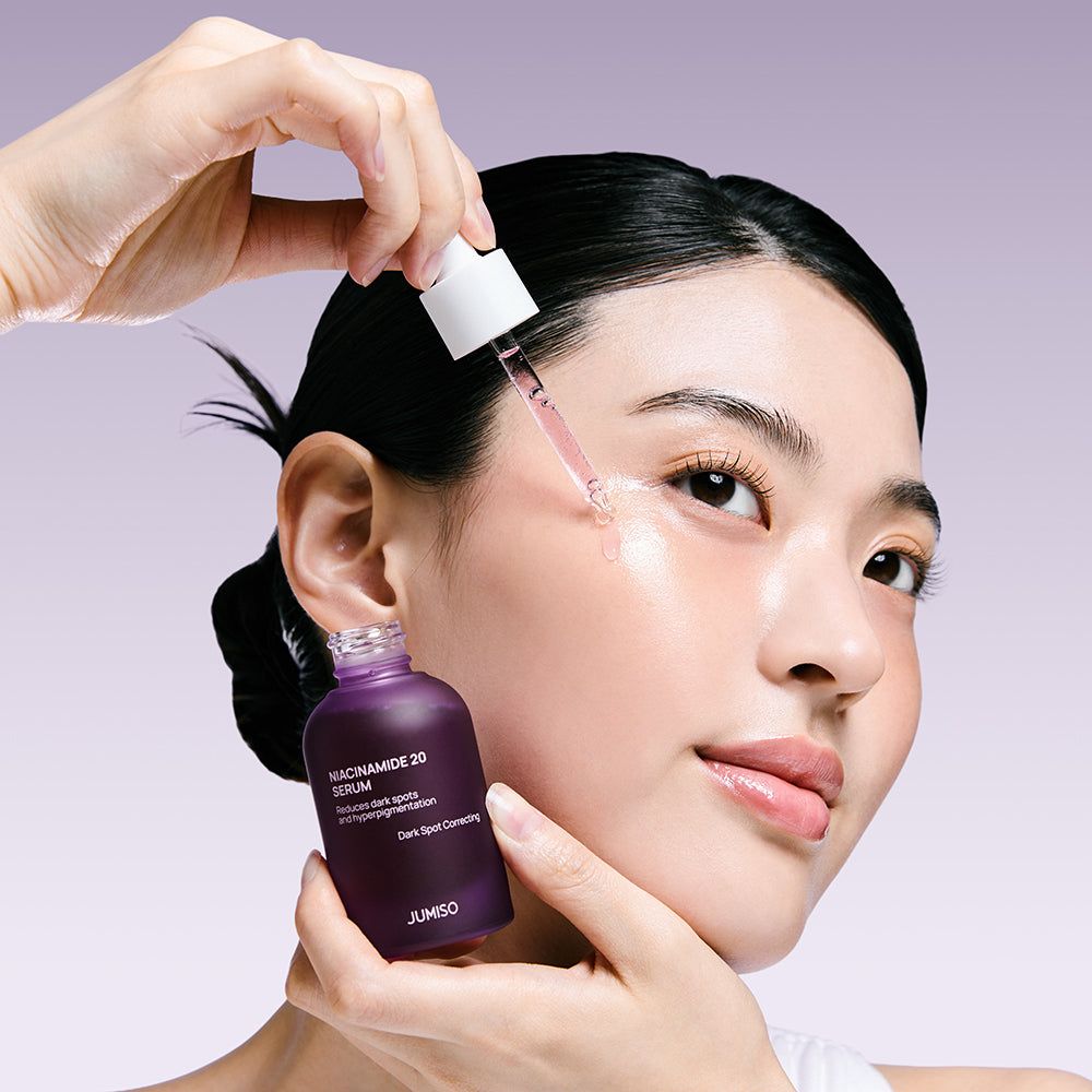 Een hand brengt Niacinamide 20 serum aan op het gezicht van een vrouw met een pipet. Donkerpaarse fles met etiket.