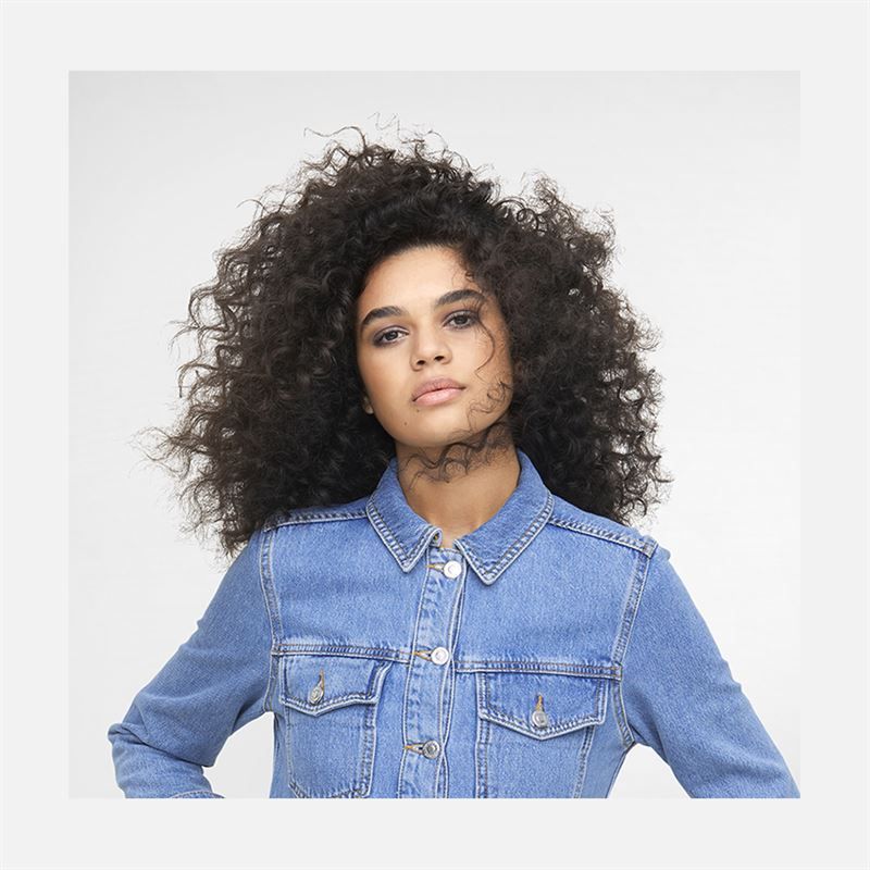 Portrait d'une femme aux cheveux bouclés. Elle porte une veste en jean. Fond clair.