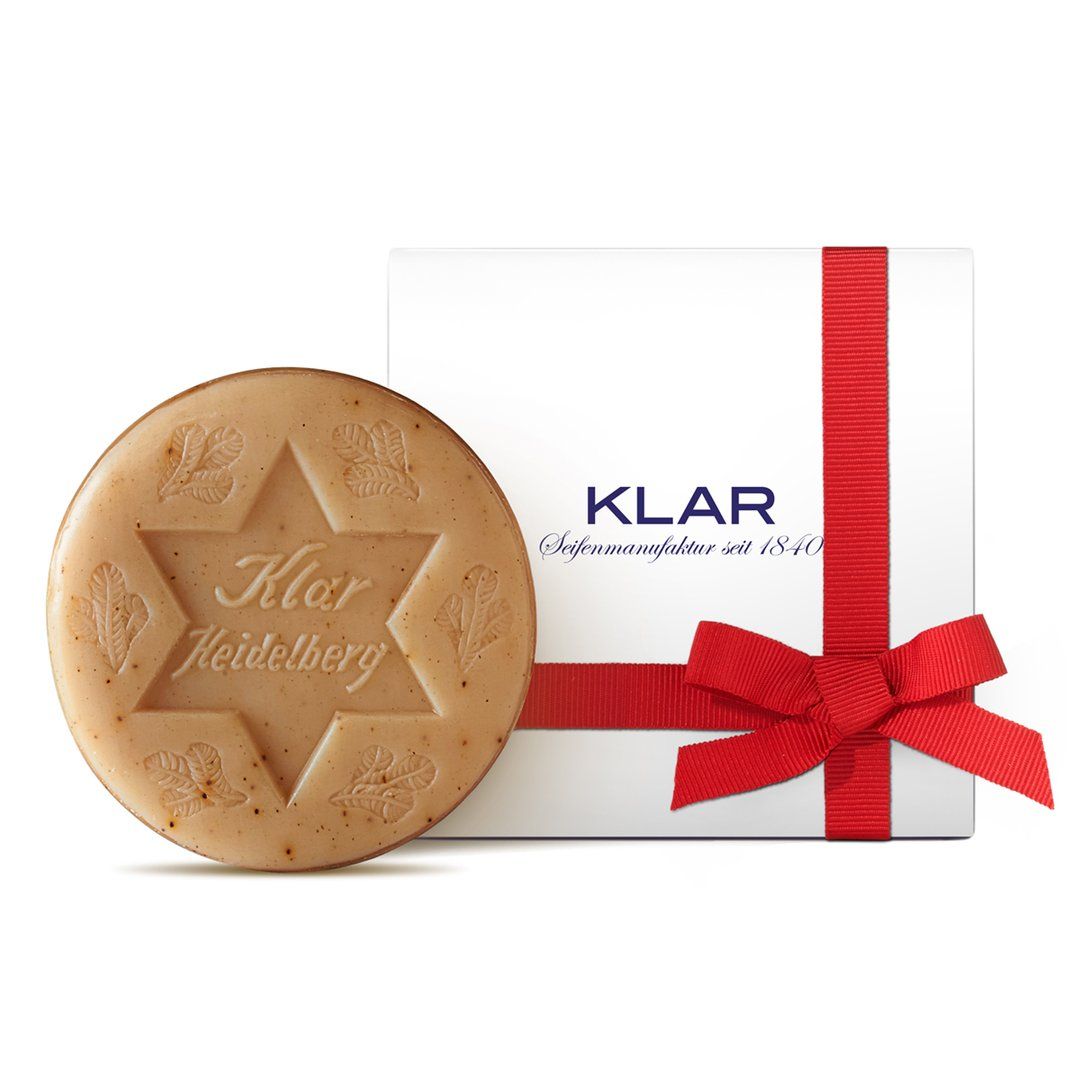 Savon rond avec motif étoile et inscription. À côté, une boîte cadeau avec un ruban rouge. Logo Klar.
