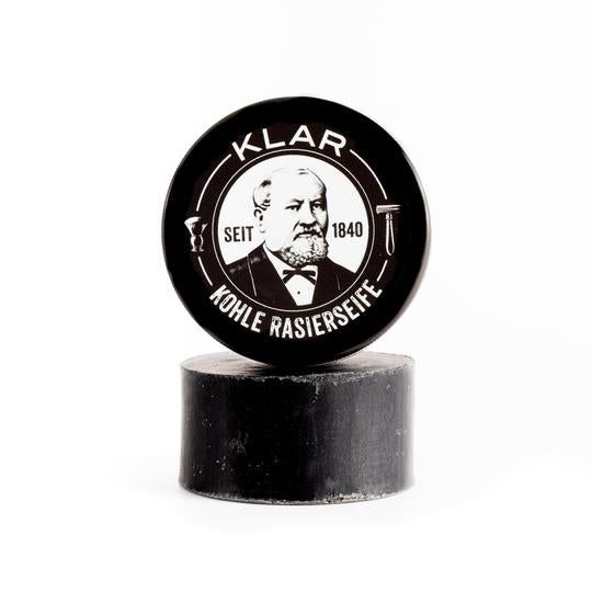 Savon à raser au charbon actif noir sur un morceau de savon noir. Couvercle avec logo et portrait. Inscription: KLAR, KOHLE RASIERSEIFE, SEIT 1840.