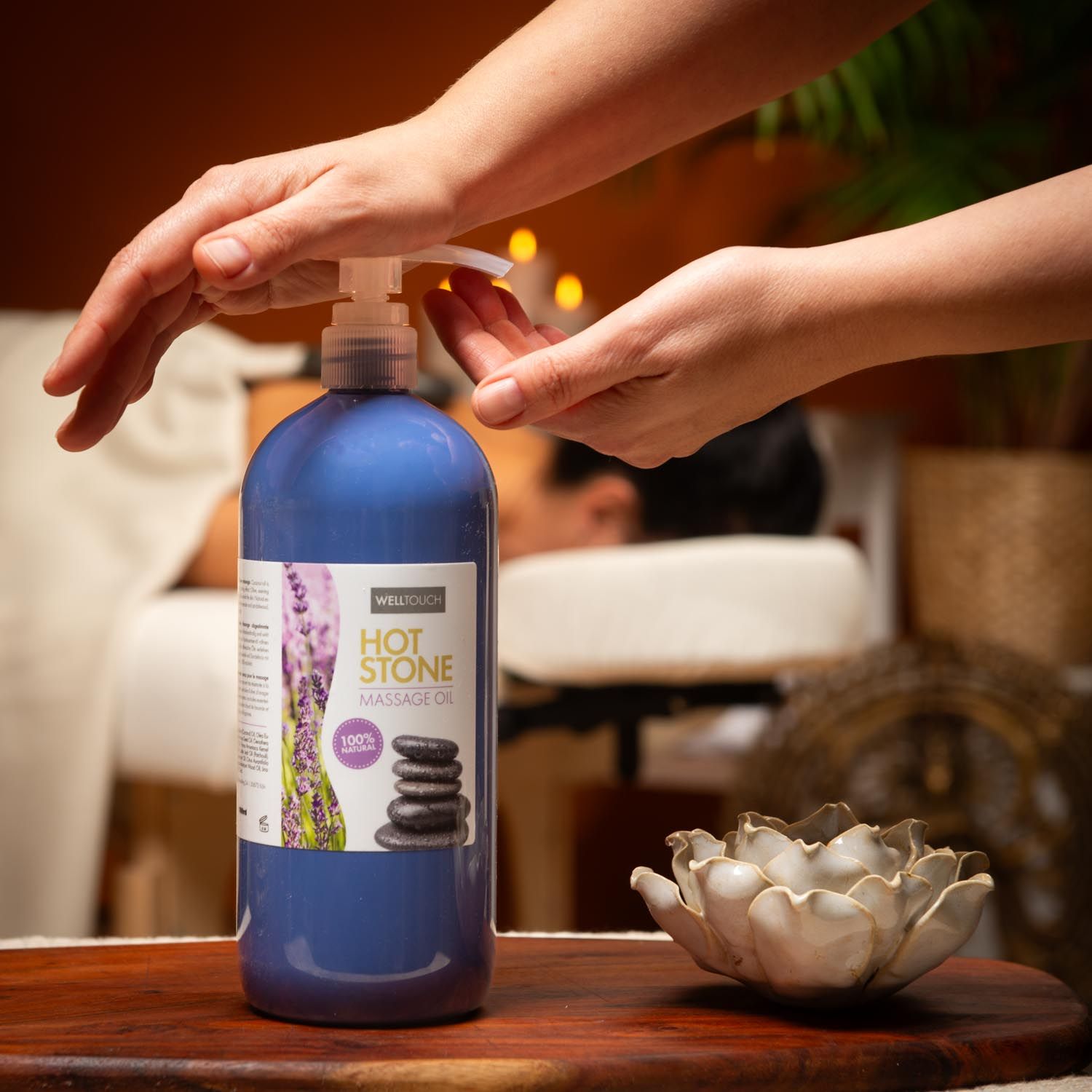 Blauwe fles Welltouch Hot Stone Massageöl met pomp. Handen drukken op de pomp. Naast lotusbloem. Achtergrond: persoon op massagetafel.