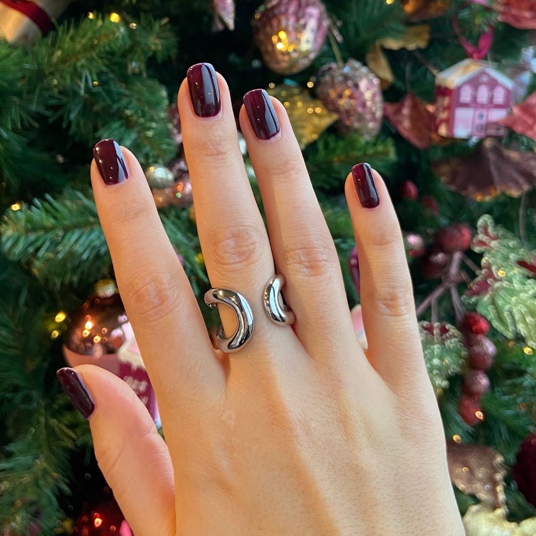 Main avec ongles vernis en rouge foncé. Les ongles sont courts et brillants. Une bague argentée est visible. Arbre de Noël en arrière-plan.