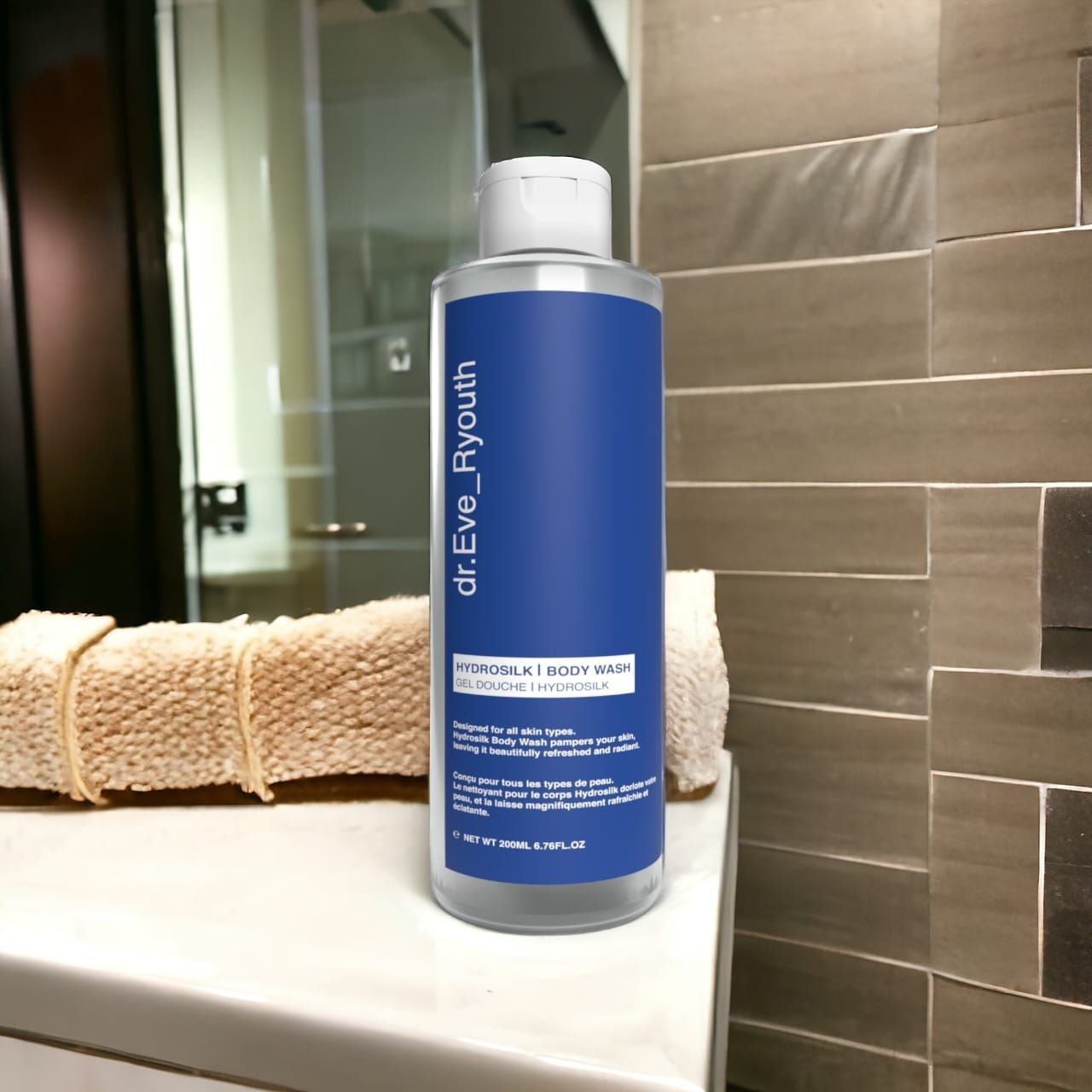 Transparante fles met blauw etiket. Opschrift: dr.Eve_Ryouth, Hydrosilk Body Wash. Op een wit oppervlak, handdoek op de achtergrond.