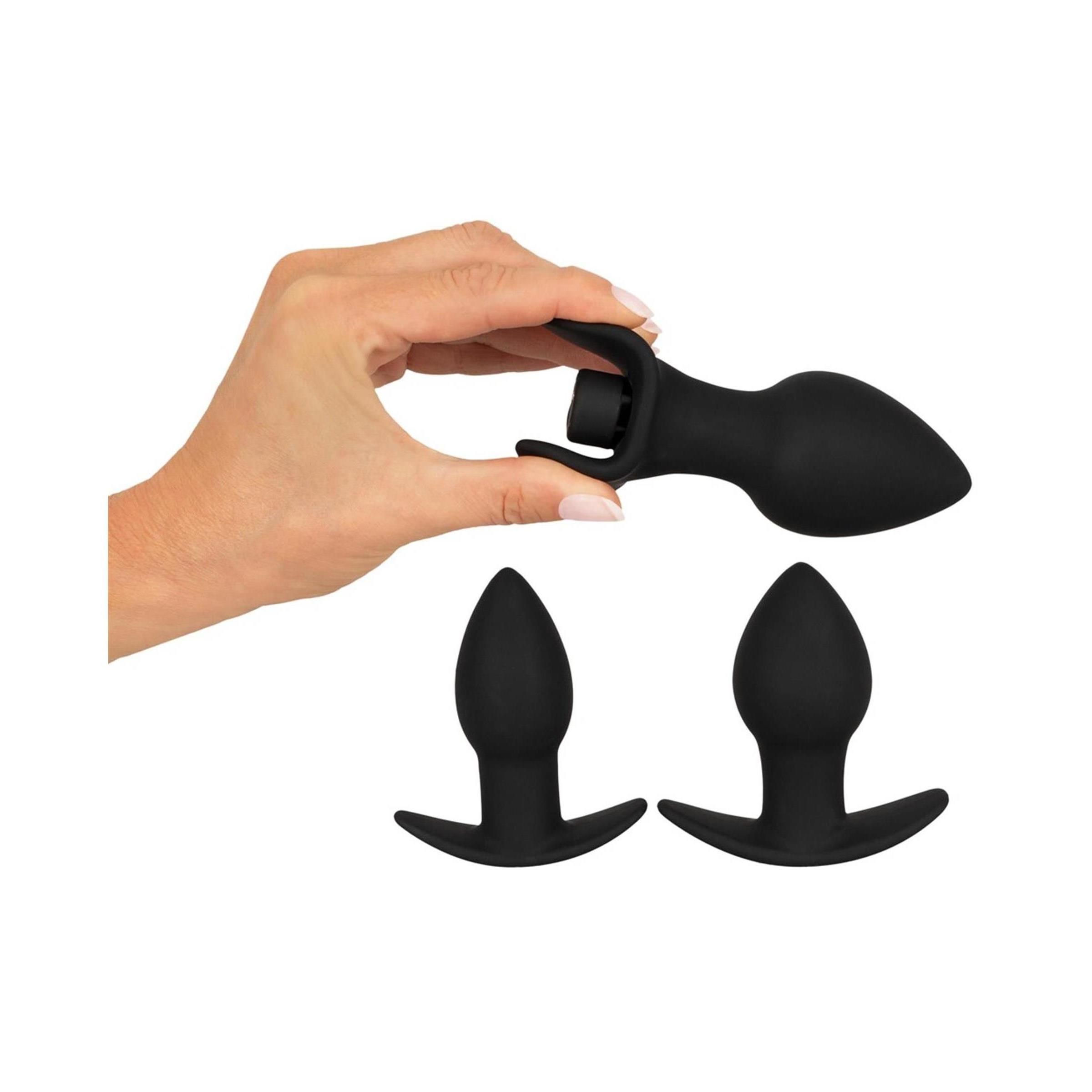 Main tenant un plug anal noir en silicone, deux autres sont posés en dessous.