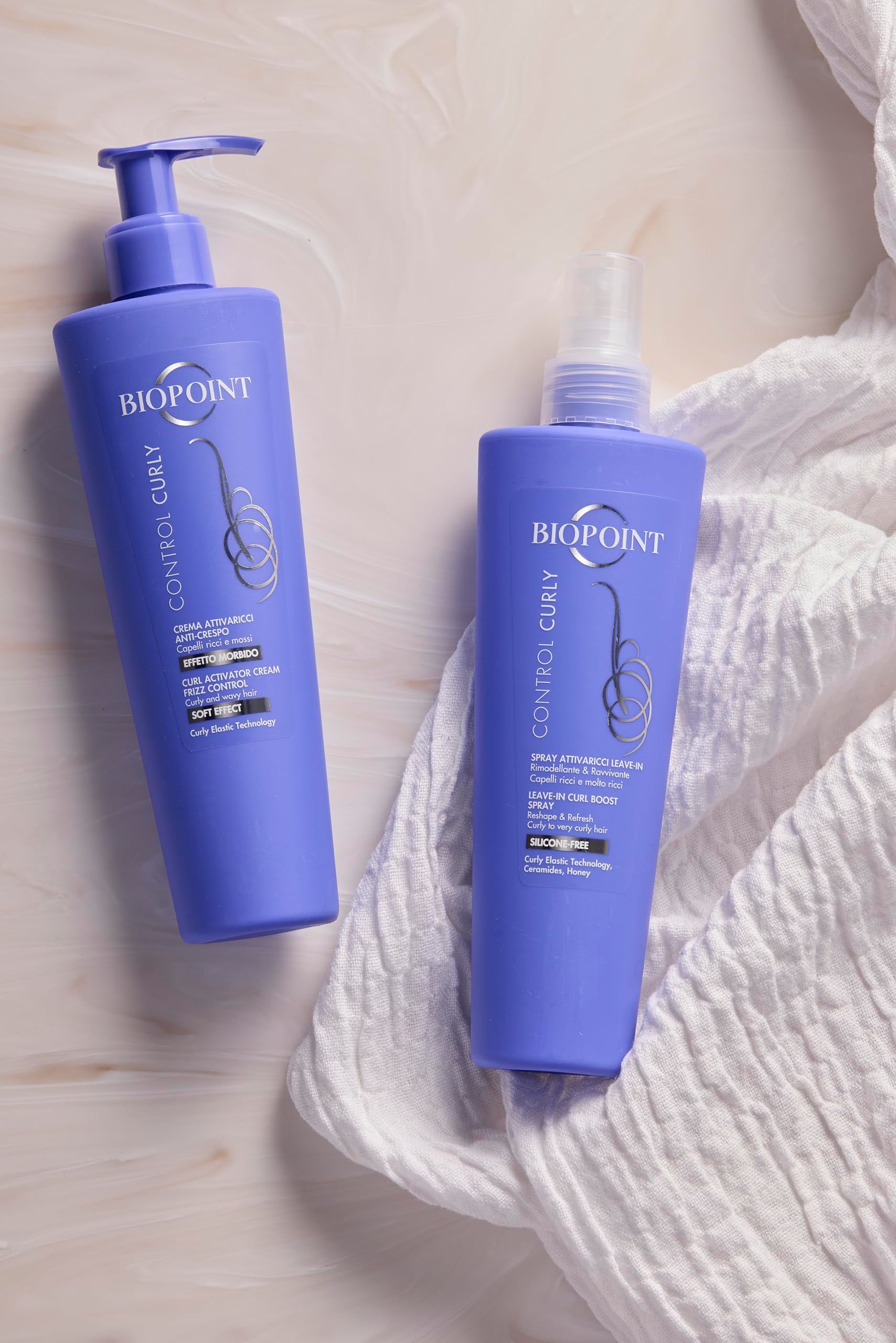 Deux produits capillaires Biopoint violets. À gauche : crème avec pompe. À droite : spray avec vaporisateur. Sur tissu blanc.