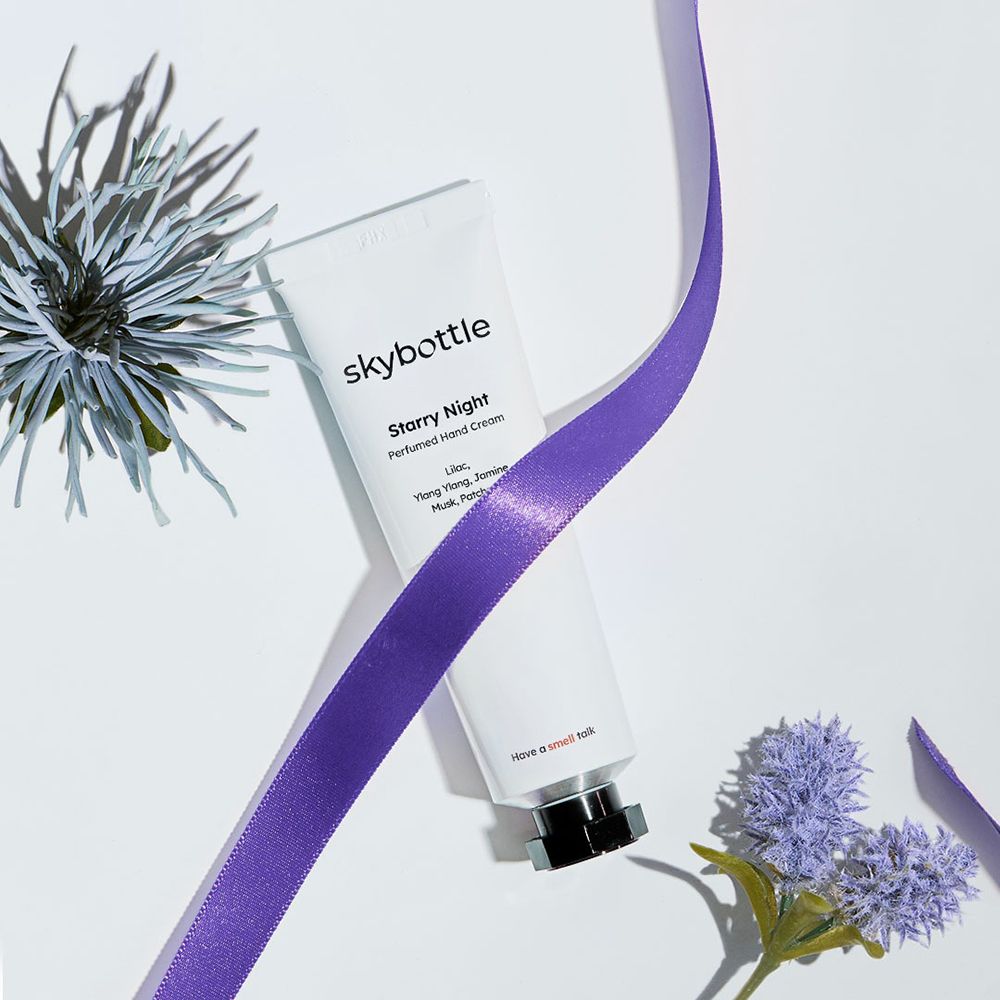 Witte tube "skybottle Starry Night Perfumed Hand Cream" met paars lint en bloemen. Zwarte dop. Op lichte achtergrond.