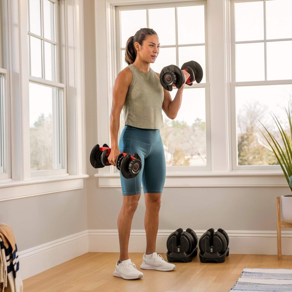 Vrouw traint met twee dumbbells. Twee andere dumbbells op de grond. Interieur met raam en tapijt.