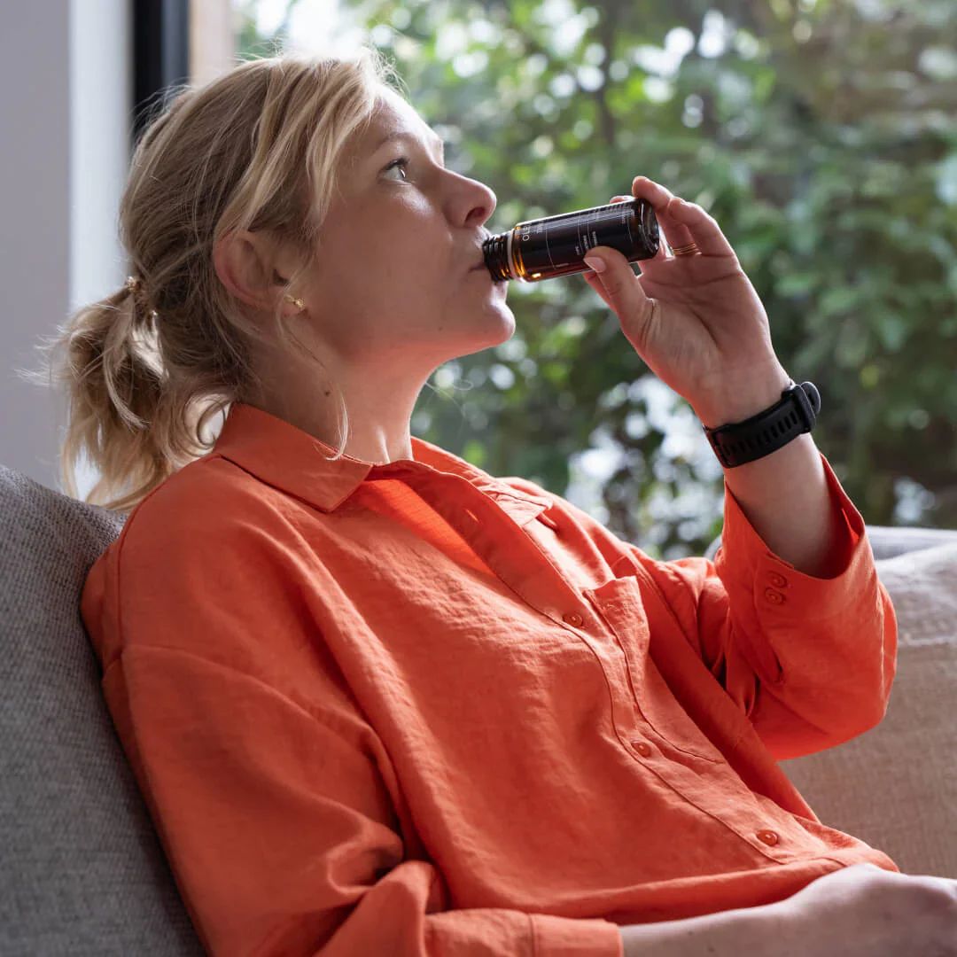 Vrouw drinkt uit flesje. OLEUS-logo en productlabel zichtbaar. Zwart flesje.