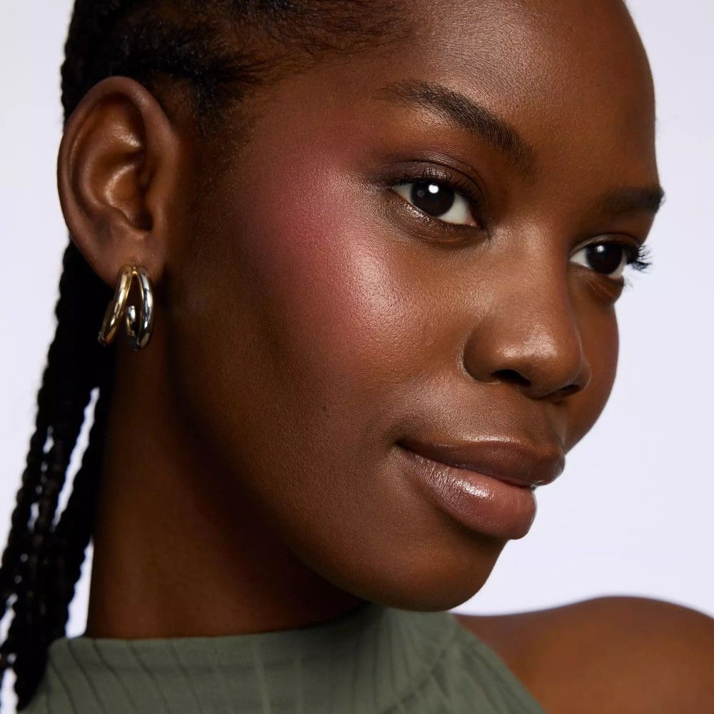 Gros plan d'une femme avec du blush rose sur les joues. Elle porte des boucles d'oreilles dorées et a la peau foncée.
