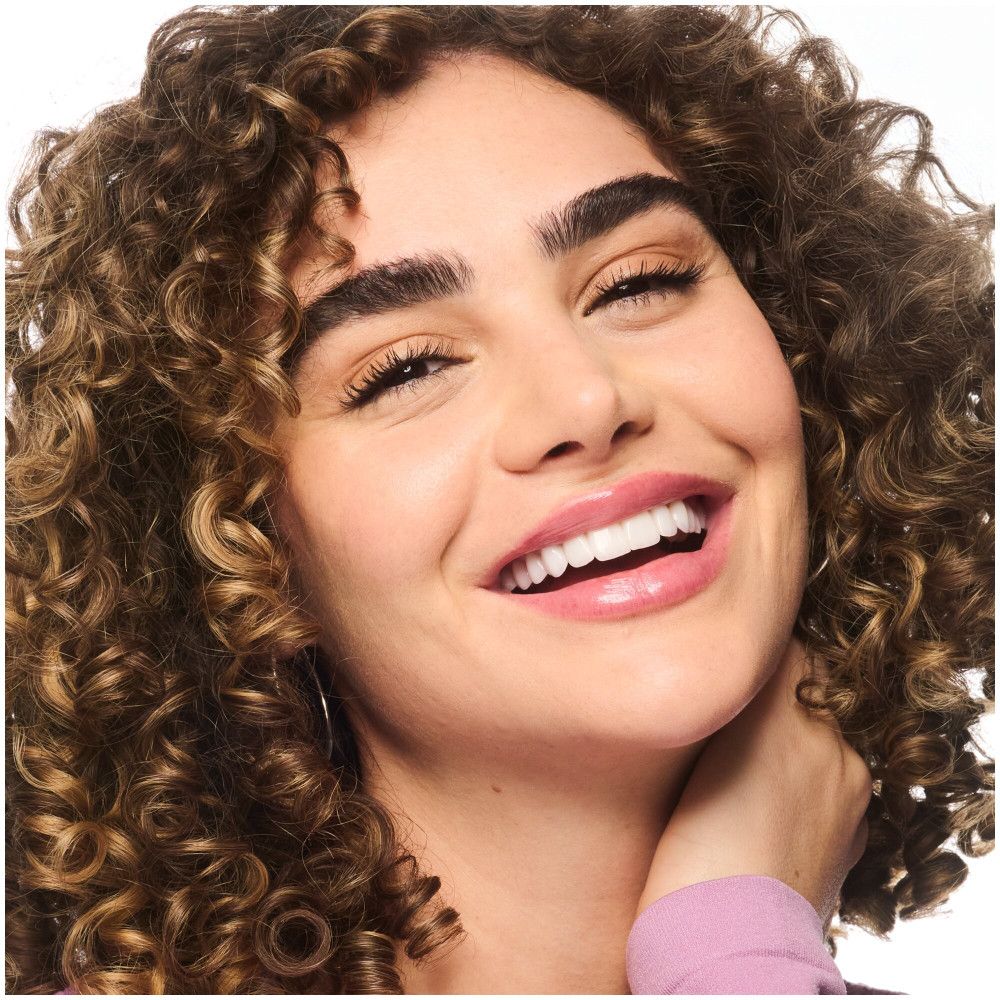 Femme bouclée souriante. Elle touche son cou. Porte un haut rose et des boucles d'oreilles.