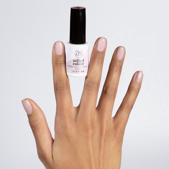 Hand met gelakte nagels en nagellakflesje. Flesje met opschrift "selfgel colour". Kleur nagellak: lichtroze.