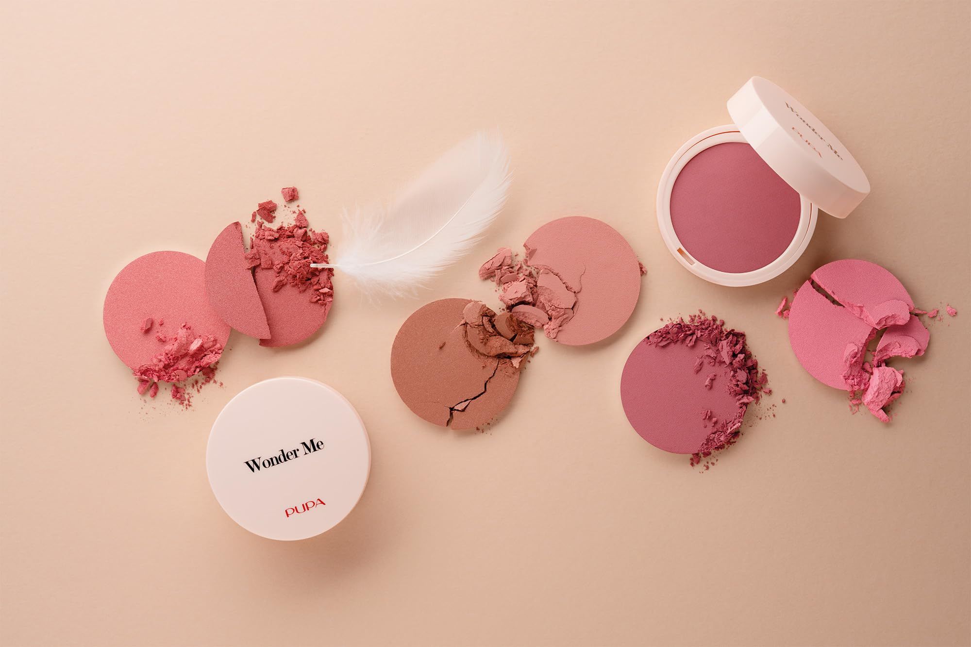 Blush-containers, gebroken poeders, veer. Verschillende roze tinten op beige achtergrond.