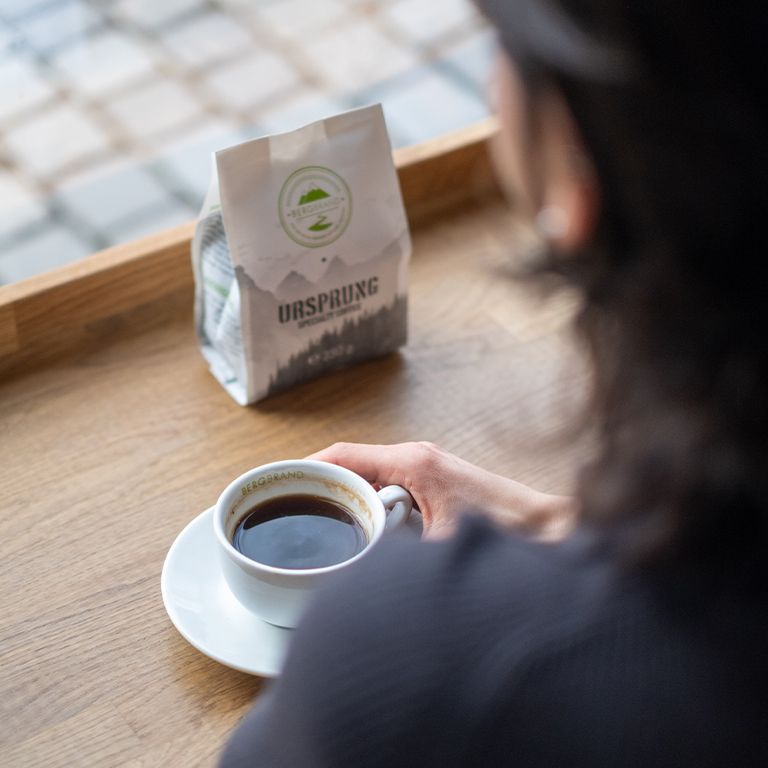 Tasse de café et paquet de grains. Paquet avec logo et texte Ursprung. Sur table en bois près fenêtre.