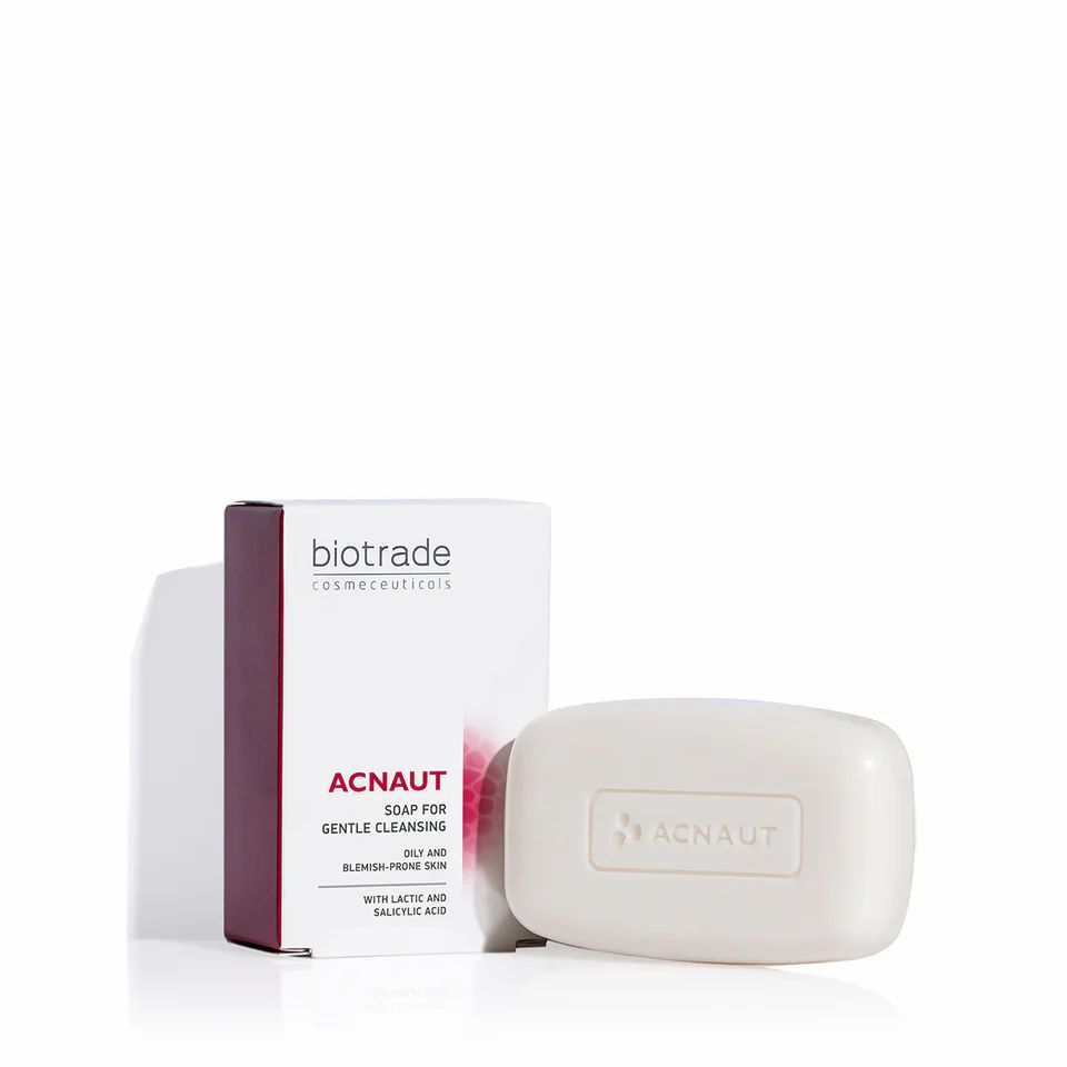 Savon et emballage. Texte : Biotrade, Acnaut Soap. Savon rectangulaire, emballage blanc avec accent rouge. Nom du produit clairement visible.